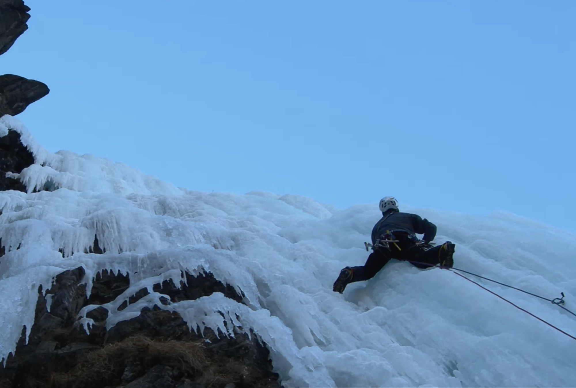 Ice climber ascending steep frozen terrain during winter conditions, viewed from below beneath a clear blue sky.