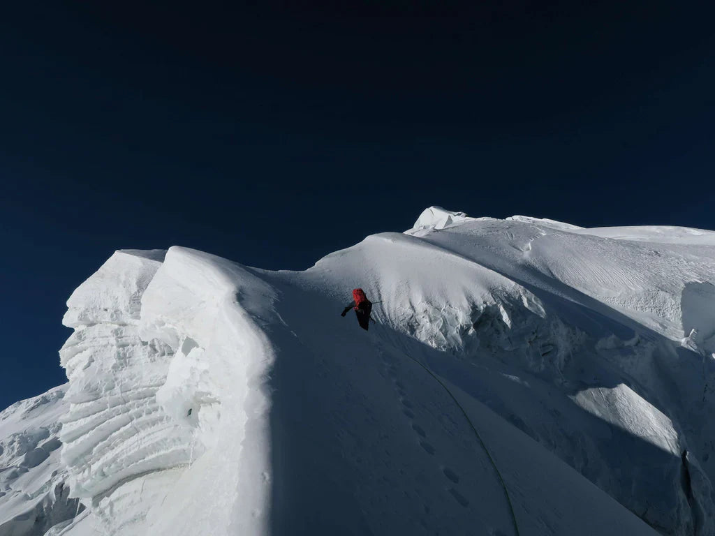 Climber following a narrow snow ridge beneath clear alpine skies.