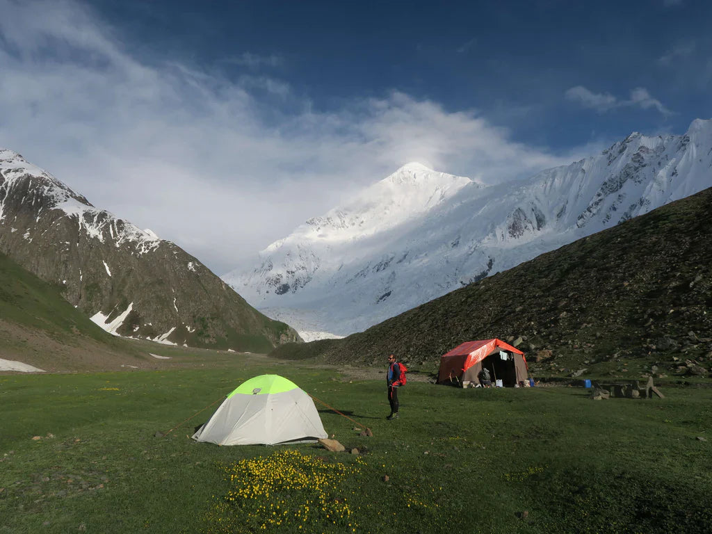 Expedition member at base camp beneath a snow-covered Karakoram peak.